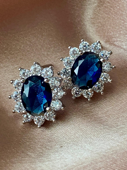 Pair of royal blue sapphire oval cut stud earrings with diamond halo, set in silver and 18K white gold, displayed on satin fabric.