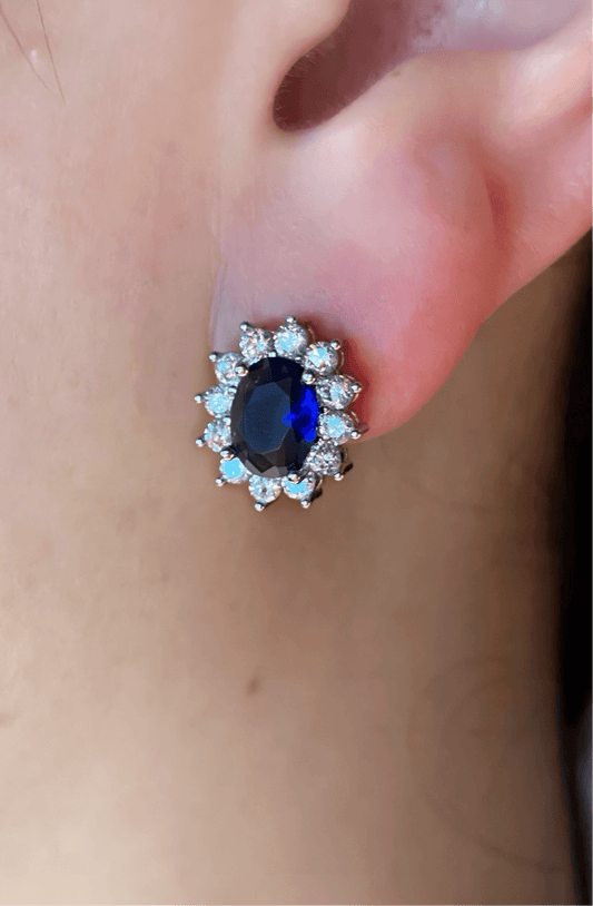 Royal blue sapphire oval cut stud earring worn on ear, surrounded by sparkling diamonds set in silver and 18K white gold.